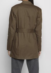 Femme portant un blazer marron ceinturé avec une fente au milieu du dos, associé à un pantalon gris ample, se tenant contre un fond blanc.