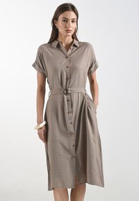 Modella che indossa un vestito beige con bottoni, stretto in vita da una cintura, con maniche arrotolate e tasche, su uno sfondo semplice.