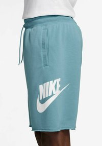 Pantaloni Nike in tessuto blu teal, con logo bianco sulla gamba sinistra, vita elasticizzata con cordino e tasche laterali.