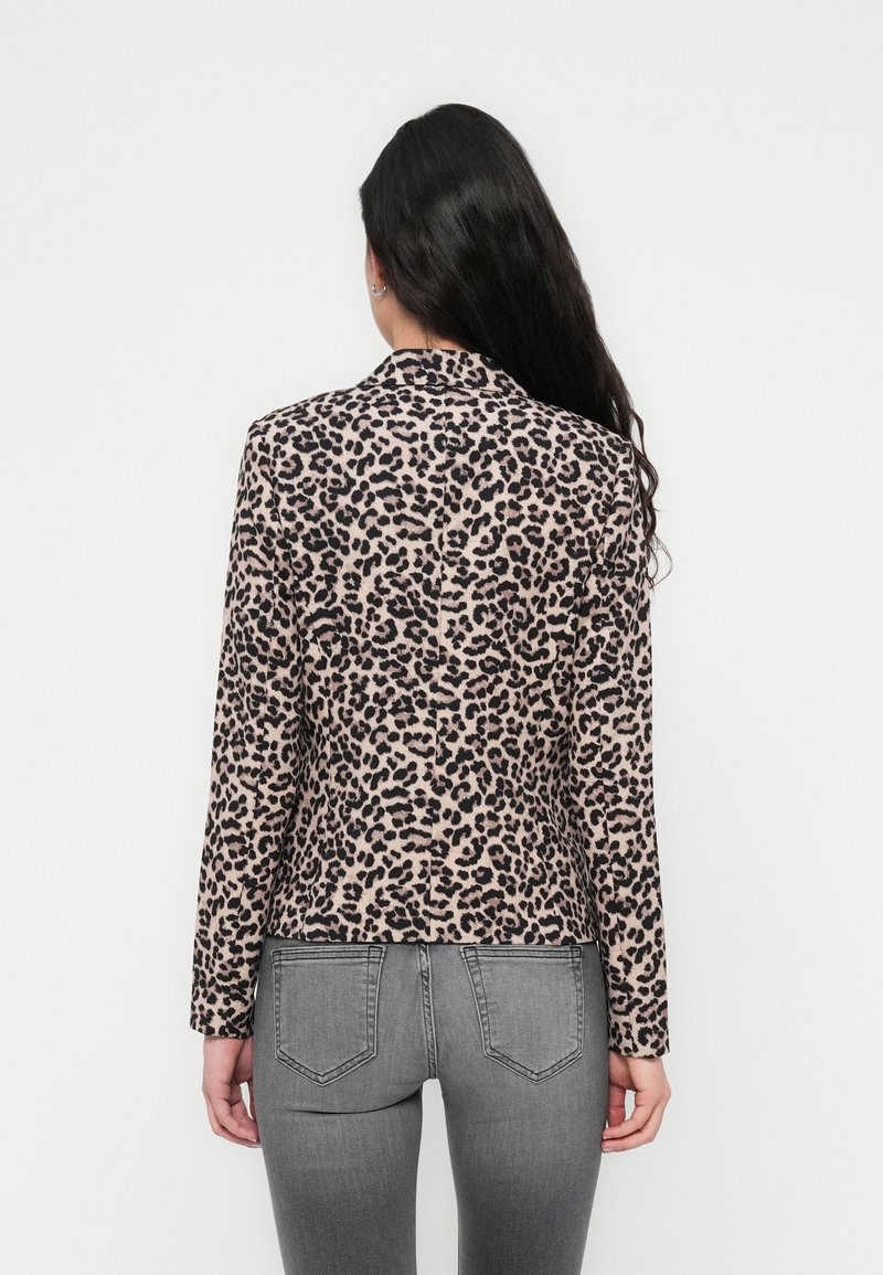 Femme aux longs cheveux foncés portant une veste beige et noire à imprimé léopard et un jean gris ajusté, vue de dos sur fond blanc.
