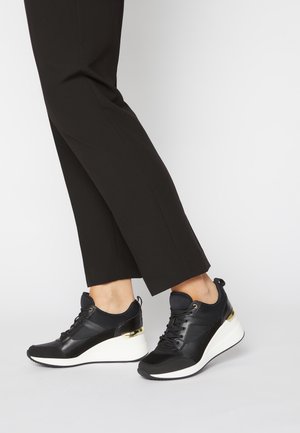 Mustat kiilasneakerit, joissa valkoiset pohjat ja kullanväriset kantakoristeet, käytetty mustujen suoralahkeisten housujen kanssa vaalealla taustalla.