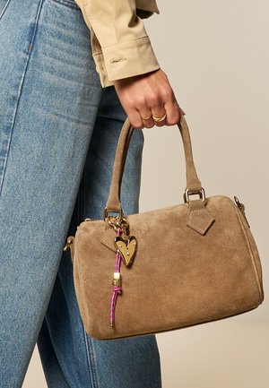 Persoon die blauwe spijkerbroek draagt en het handvat vasthoudt van een lichtbruine suède handtas met gouden hartbedel en roze koorddetail.