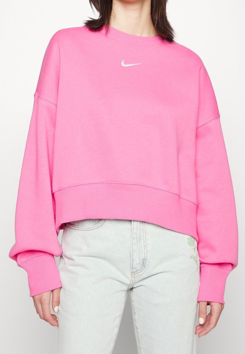 Žena v oversized jasně růžovém svetru Nike a světlých modrých džínách s vysokým pasem, stojící proti jednoduchému bílému pozadí.