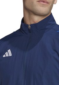 Leichte navyblaue Jacke mit hohem Kragen, durchgehendem Reißverschluss und weißem Logo. Glattes, strukturiertes Material mit kontrastierenden blauen Akzenten.