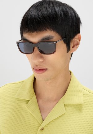 Giovane uomo con capelli corti e neri che indossa occhiali da sole rettangolari scuri e una camicia texturizzata giallo chiaro con colletto.