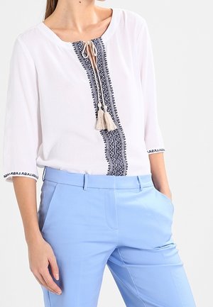 Vrouw die een witte blouse draagt met blauw geborduurde patronen en kwastjes, gecombineerd met lichtblauwe op maat gemaakte broek, handen in de zakken.