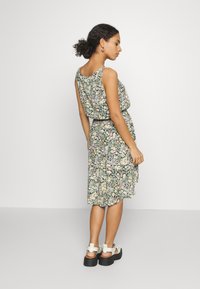 Vestido floral em verde, com um design solto e de mangas curtas, cintura elástica e saia até ao joelho, combinado com sandálias de plataforma pretas.