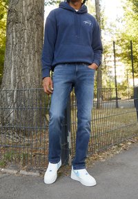 Navy modrá bavlněná mikina s logem, světlé modré denimové džíny a bílé tenisky s modrými akcenty, stojící u stromu a plotu.