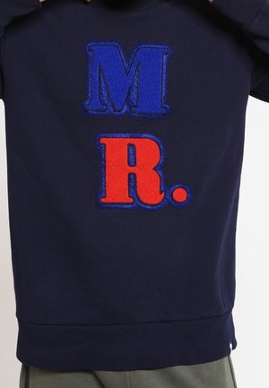 Pull bleu marine avec de grandes lettres en relief "M" en bleu et "R." en rouge dans le dos.