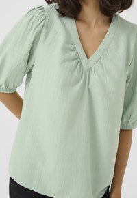 Femme portant un haut virement en V texturé de couleur vert clair avec des manches mi-longues bouffantes et des poignets élastiqués, associé à un bas noir.