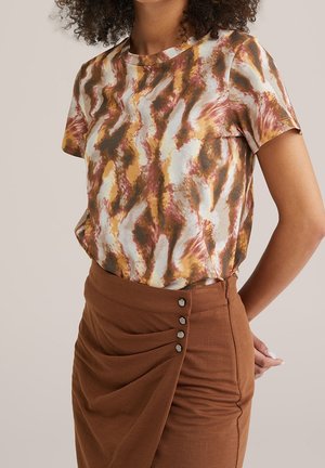 Vrouw draagt een shirt met korte mouwen met een abstract bruin, beige en roestkleurig patroon, ingestopt in een bruine rok met zijknopen en plooien.
