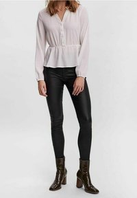 Weiße Bluse mit langen Ärmeln, Knopfausschnitt in V-Form und Taillenspiegel, kombiniert mit schwarzer enger Hose und glänzenden braunen Stiefeletten.