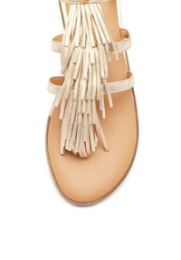 Sandalo piatto beige con molteplici fasce sottili e una frangia decorativa realizzata in materiale testurizzato, con una suola liscia di colore chiaro.