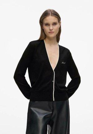 Vrouw staand met handen achter rug, draagt een zwart gebreid vest met knopen en witte bies en een zwarte leren broek tegen een lichte effen achtergrond.