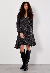 Schwarzes Kleid mit Leopardenmuster, langen Ärmeln und gerafftem Bund. Getragen mit kniehohen schwarzen Lederstiefeln.