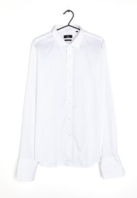 Camisa blanca de manga larga, abotonada, hecha de tejido liso, con un cuello clásico, botones en el frente y puños anchos. Cuelga de un gancho negro.