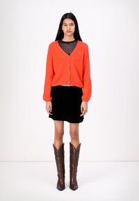 Cardigan tricoté orange à manches longues et boutons en perle, associé à une jupe en velours noir et des bottes cowboy brodées marron.