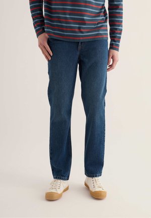 Person iført blå denim jeans, hvide sneakers med gummisåler og en langærmet skjorte med blå og røde vandrette striber.