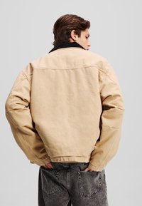 Beige bomulljakke med svart krage, avslappet passform og synlig søm. Brukt med mørkegrå jeans med baklommer.