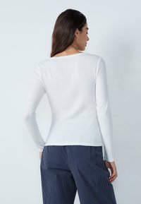 Maglietta bianca a maniche lunghe con un ampio scollo, abbinata a pantaloni blu a righe sottili. I tessuti hanno una superficie liscia e una leggera lucentezza.