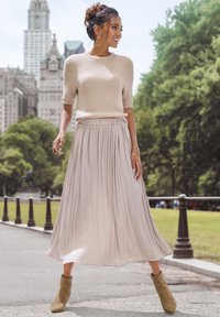 Maglione beige a maniche corte in maglia con una consistenza morbida abbinato a una gonna maxi plissettata di un leggero beige. La modella indossa stivaletti alla caviglia verde oliva.