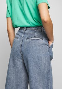 Vaaleansiniset farkut, joissa on korkea vyötärö, leveät lahkeet ja kaksi takataskua, yhdistettynä lyhythihaiseen vihreään T-paitaan.