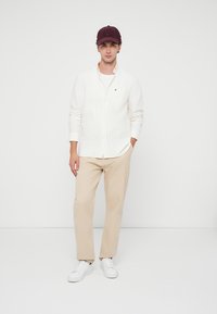 Giovane uomo che indossa una camicia bianca con bottoni, pantaloni beige, sneakers bianche e un cappellino da baseball bordeaux, in piedi di fronte a uno sfondo bianco uniforme.