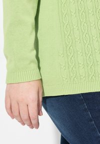 Vaaleanvihreä villapaita, jossa on keskellä kabelikuvioinen muotoilu, ribbireunat hihoissa ja helmassa, yhdistettynä tummansinihin farkkuihin.