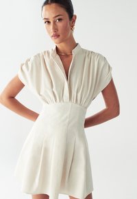 Lyhythihainen, beige mekko, jossa on V-pääntie, rypytetty vyötärö ja liehuva hame. Valmistettu kevyestä kankaasta, jossa on hienovarainen rakenne ja laskokset.