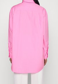 Langärmeliges Hemd aus leuchtend rosa Baumwolle mit durchgehender Knopfleiste am Rücken, Kragen und aufgekrempelten Manschetten. Der gerade geschnittene Saum reicht etwas unter die Taille.