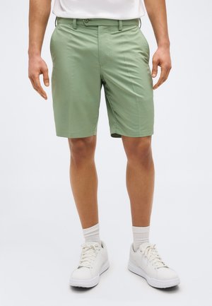 Homme portant un short vert clair jusqu'aux genoux, des chaussettes blanches, des baskets blanches et une chemise blanche, debout devant un fond uni.