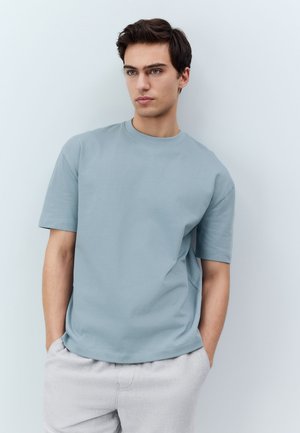 Giovane uomo con capelli scuri che indossa una camicia azzurro chiaro a maniche corte e pantaloni grigio chiaro, in piedi contro un muro bianco semplice, con le mani in tasca.