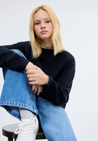 Mornarska majica s dolgimi rokavi, modri denim jeans in bele teniske. Svetlo blond lasje obdajajo obraz na običajni ozadju.