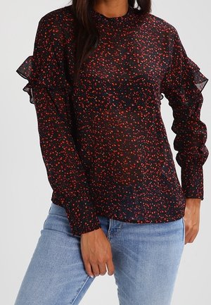 Woman wears a sheer black blouse with red and orange speckles, ruffled long sleeves, and light blue jeans.