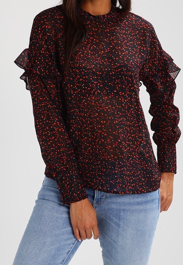 Femme porte un chemisier noir transparent avec des taches rouges et orange, des manches longues à volants et un jean bleu clair.