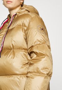 Złota kurtka puchowa z kapturem, z zamkiem błyskawicznym w czerwono-biało-niebieskie akcenty oraz naszywką z logo na lewej rękawie. Gładka tekstura.