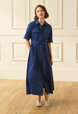 Frau steht drinnen und trägt ein knielanges blaues Hemdblusenkleid mit kurzen Ärmeln, einem gebundenen Taillengürtel und weißen gemusterten Slip-on-Schuhen.