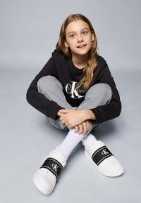 Biele tenisky Calvin Klein s širokým čiernym pásom s logom. Model má na sebe sivé teplákové nohavice a čiernu mikinu. Pozadie je svetlomodré.