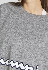 Grå sweatshirt med texturerad tyg, rund nacke och framträdande "Champion"-logotyp i svart och vitt längs nederkanten.