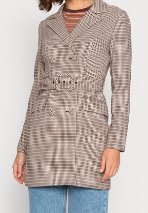 Femme portant un blazer ceinturé à double boutonnage pied-de-poule sur un haut rayé marron et un jean bleu clair, debout avec une main détendue.