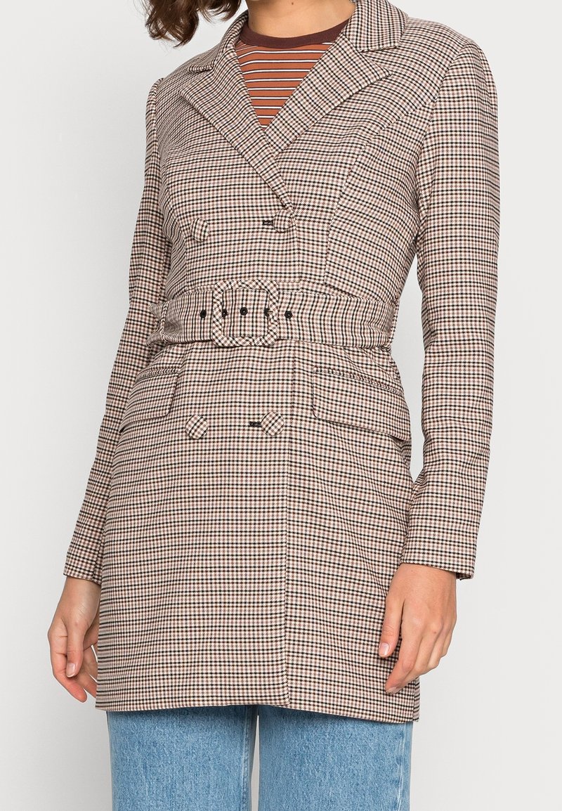 Femme portant un blazer ceinturé à double boutonnage pied-de-poule sur un haut rayé marron et un jean bleu clair, debout avec une main détendue.