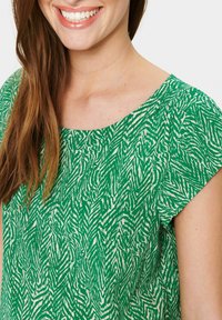 Blusa verde a fantasia con scollatura rotonda e maniche a cappuccio, caratterizzata da un design textured di linee zigzag bianche.
