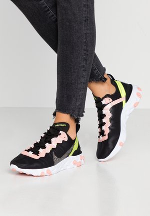 Stopy w czarnych butach Nike React z różowymi i neonowo-żółtymi akcentami oraz białymi podeszwami, w połączeniu z postrzępionymi czarnymi wąskimi dżinsami.