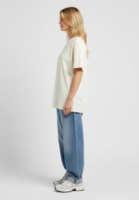 Krémová oversized tričko, krátké rukávy, spárované se světle modrými volnými džíny a bílými teniskami se šedými akcenty. Jednoduchý design.
