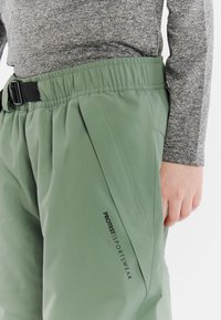 Groene waterdichte broek met een elastische tailleband en zwarte gesp, voorzien van zijzakken en het logo "PROTEST SPORTSWEAR" in het zwart.