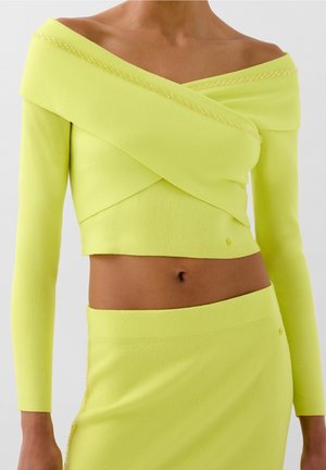 Kobieta ubrana w jasny żółty, odkryte ramiona, długi rękaw, krótki top oraz dopasowaną spódnicę z wysokim stanem na neutralnym tle.