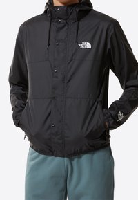 Person som bär en svart North Face-hoodie med tryckknappar och turkosa byxor, med händerna i jackfickorna, mot en vit bakgrund.