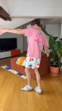 Noormees värvilistes lühikestes pükstes ja roosas särgis sirutab käsi moodsas elutoas, kus on puidust põrand ja suur roheline taim.