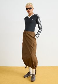 Blusa preta de manga longa com gola branca e logo da Adidas; saia cargo castanha até ao tornozelo com cordão na bainha; sapatilhas cinzas e castanhas.