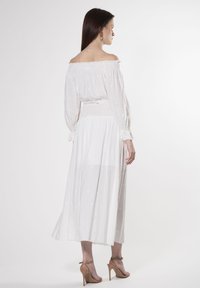 Hvid off-shoulder kjole med lange ærmer, sammensat talje og broderede detaljer. Fløjlende nederdel der når til midten af læggen; parret med nude hæle.
